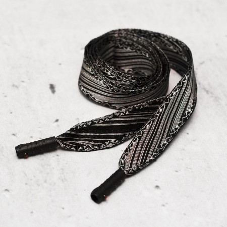 Ozdobny sznurek wykonany ze srerno-czarnej taśmy żakardowej, zakończony z obu stron ozdobną skuwką.
